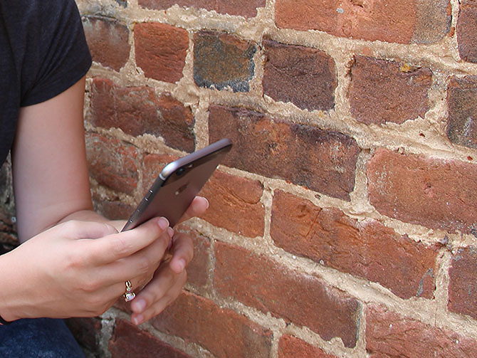 A woman checks her phone while sitting beside a brick wall.