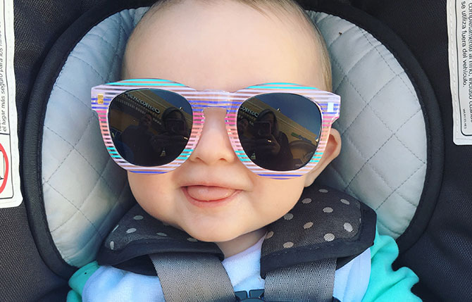 A cute baby girl grins from her car seat in big sunglasses.