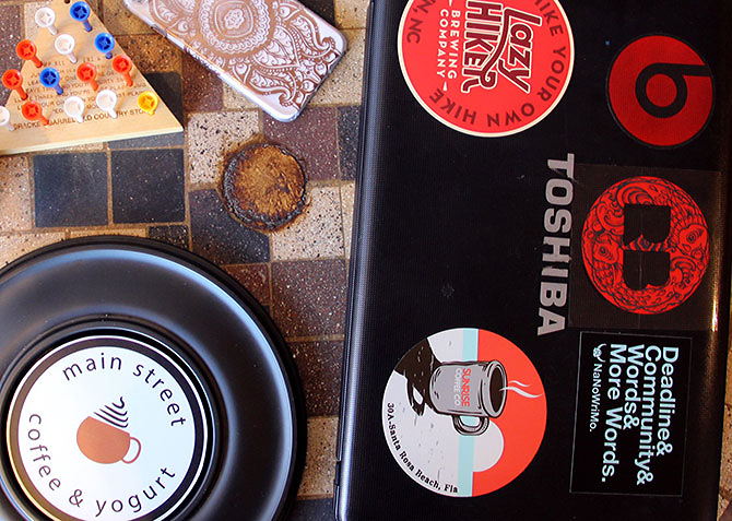 A stickered laptop, phone, and coffee cup coaster on a tile table in a coffee shop