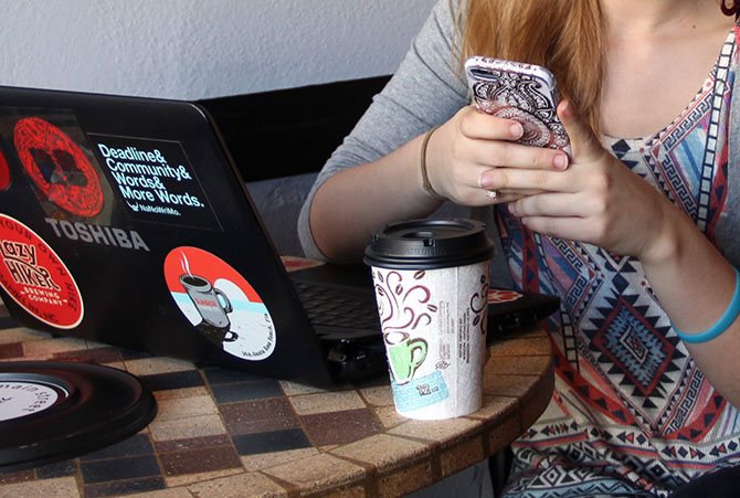 A girl checks her phone while using her laptop in a coffee shop.