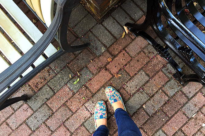 Blue shoes walk up to two iron benches on a brick sidewalk.
