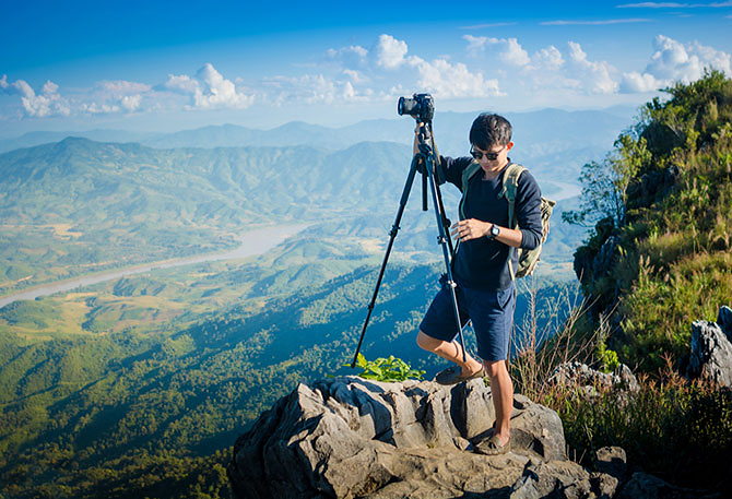Young man carries his camera and tripod up a rock, with mountains stretching into the distance behind.]