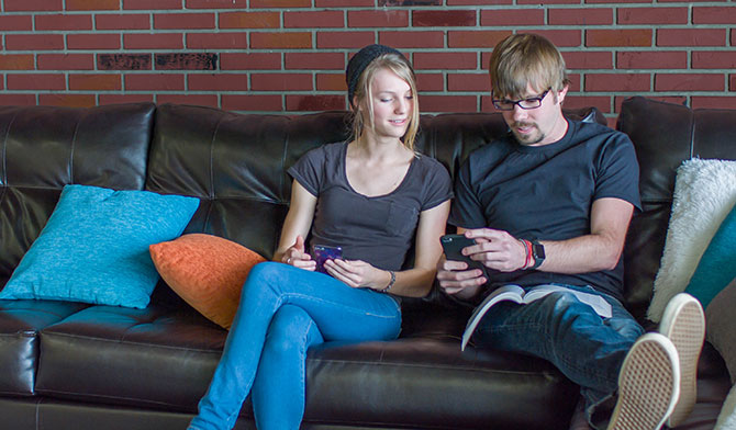 Two young adults talking and looking at their phones on a couch.