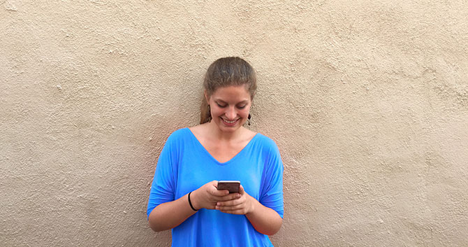 Girl in a blue shirt leans against a wall and grins at phone.