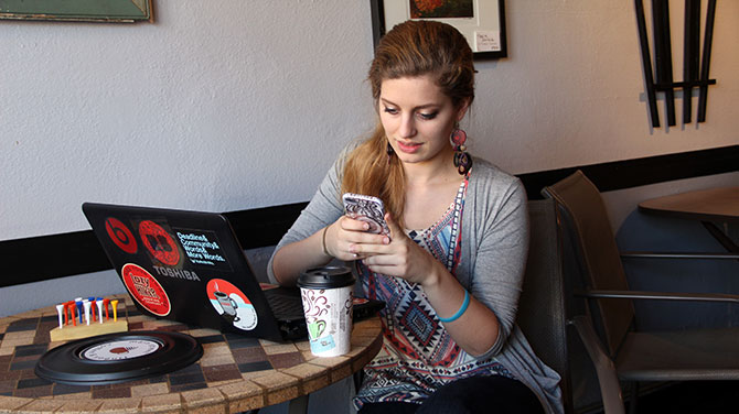 A woman smiles while using her phone and laptop in a coffee shop.