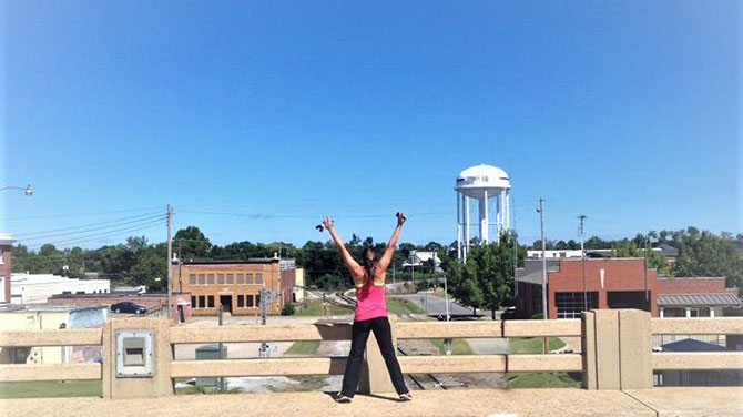 A woman stands on a bridge with her hands raised high, overlooking a small town below.