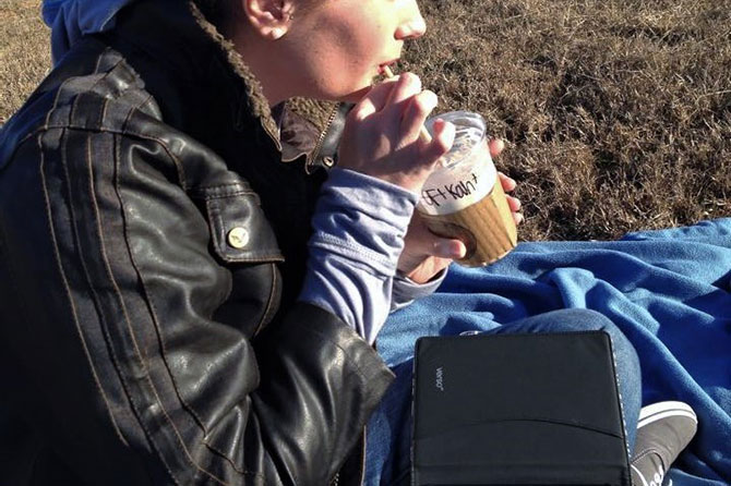 A woman sips coffee on a blanket outside with her tablet in her lap.