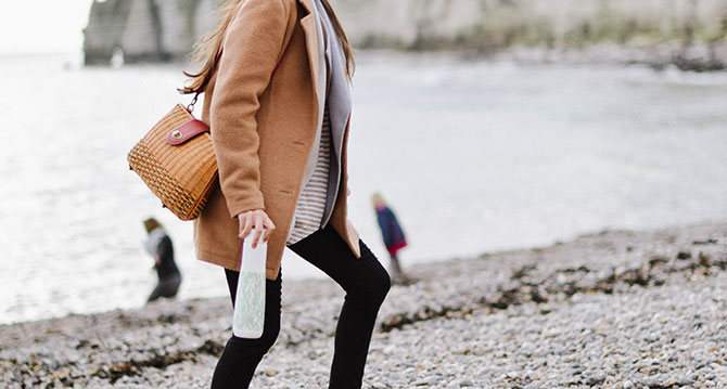 A young woman walking across a pebbly beach holding a sustainable water bottle.