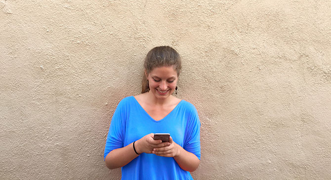 A young woman leaning against a wall and smiling at her phone.